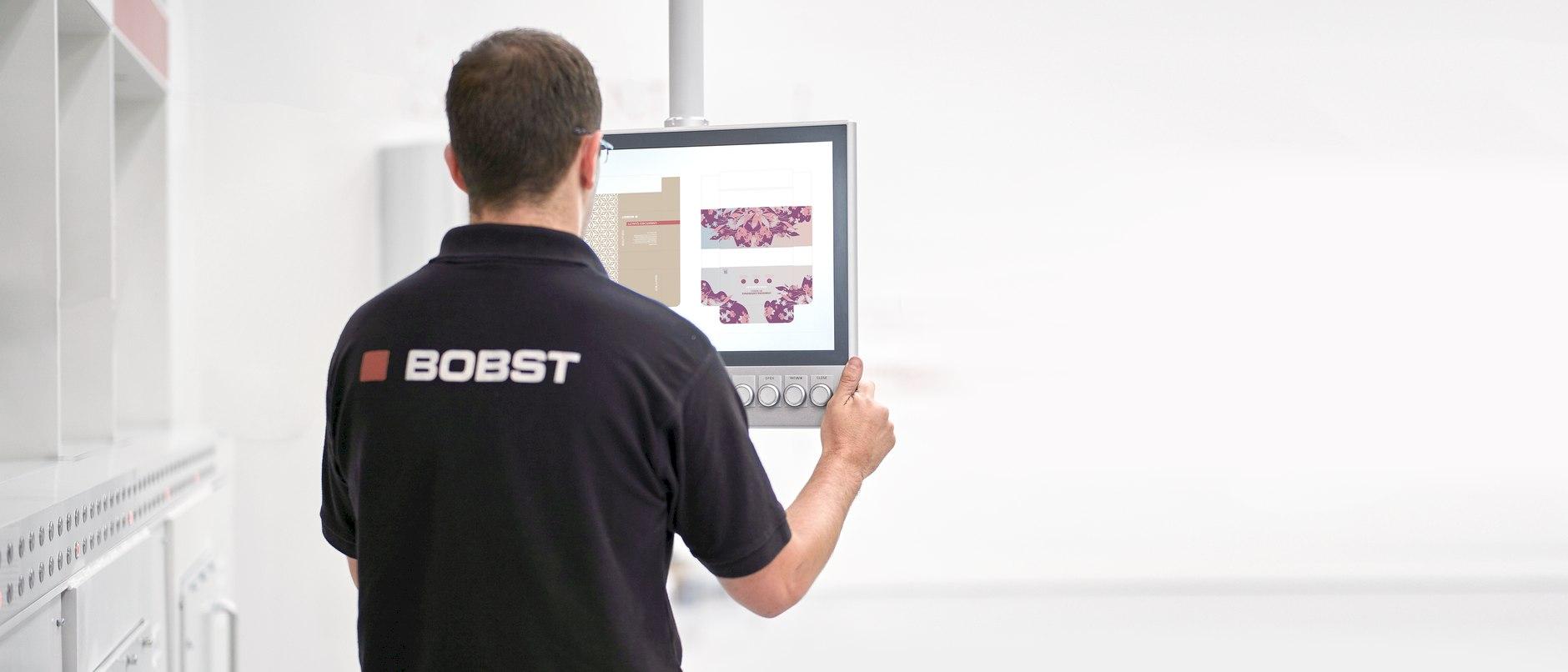 A man wearing a BOBST shirt standing in front of an industrial control display